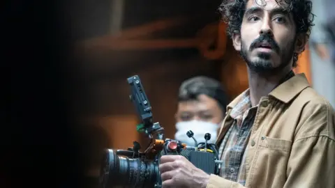 Universal Dev Patel on set for Monkey Man. Dev is a 33-year-old British Asian man with curly dark hair, brown eyes and a short beard and moustache. He holds a professional camera and looks over his left shoulder. He is wearing a camel coloured over shirt with a plaid brown and orange shirt. He’s pictured inside with a member of crew in the background wearing a face mask