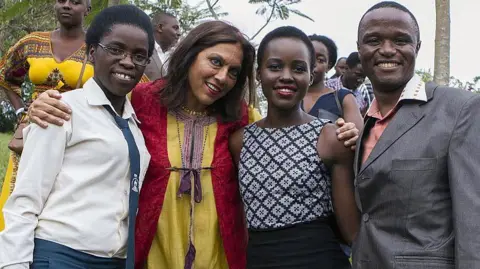 AFP/Getty Images Four people pose for a photo in Kampala in 2015. Ugandan chess champion Phiona Mutesi is on the left, wearing a beige shirt and green tie. Next to her is film-maker Mira Nair in a red and yellow kaftan, who has her arms around Phiona and actress Lupita Nyong'o, in a black and white sleeveless outfit with Robert Katende ,in a grey jacket and open-necked pink salmon shirt, standing on the right.