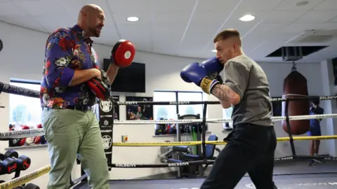 Rays of Sunshine A young short haired man wearing boxing gloves is holding them close to his face before he attempts to throw a punch at Tyson Fury who is holding two hook and jab pads