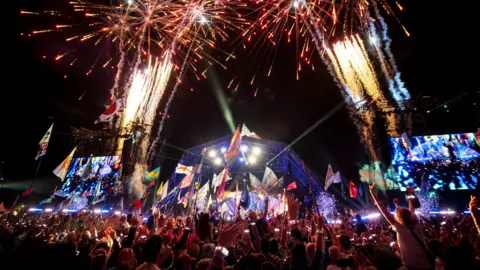 Pyramid Stage at Glastonbury during Coldplay's headline set. It is dark but the sky is lit up with a firework display and the large crowd is waving flags with their arms up in the air. On either side of the triangular stage are electronic screens which are glowing with UV light special effects. 