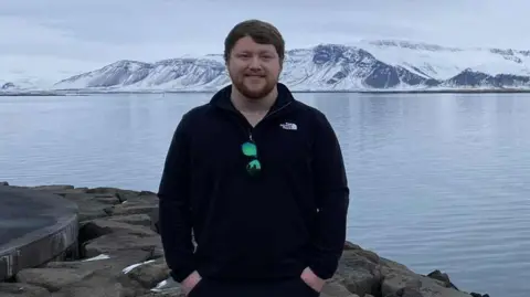 IOM CONSTABULARY Jack poses for a photo. He's wearing dark clothes, and has dark hair and a ginger beard. He has his hands in his pockets and is smiling. There is lake and snow covered hills behind him.