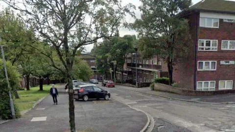 Google A man walks past some parked cars on a large pavement across a road from a complex of flats. There are trees lining each side of the road.