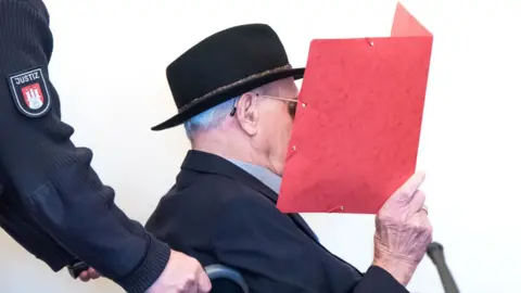 AFP Mr Dey is seated in a wheelchair, wearing a black hat and sunglasses, as he holds a red folder up in front of his face