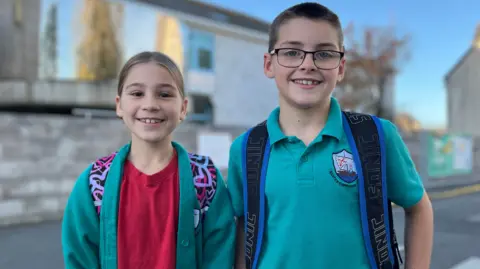 Inara and Sebastian - On the left is a young girl with blonde hair smiling at the camera. She's wearing a turquoise jumper and a red shirt. She has a patterned back back on her back and you can see the straps. On the right is a taller boy with black hair, black glasses and a turquoise polo shirt. He has a backpack on with blue and back straps. 