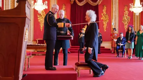 PA Media King Charles lays a sword on the shoulder of Brian May while knighting him at Buckingham Palace