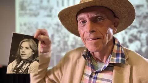 Bruce Bayley holds up an old photograph of himself and smiles to the camera