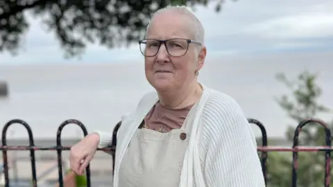Sally is leaning on railings with the sea behind her. She is wearing a cream cardigan, brown t-shirt and light brown dungarees. She has short white hair and glasses.