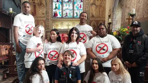 Wycombe Youth Action Group photo inside a large historic church with ornate painted walls, a vaulted wooden ceiling and a stained-glass window behind the group. Eleven people are gathered around a uniformed police officer kneeling at the front. Most of the group are wearing matching white T-shirts featuring a red crossed-out knife symbol and the text “NO KNIVES”. The group is positioned on a red carpet in front of the altar, with floral arrangements, religious artwork and church furnishings visible in the background. A person in dark clothing stands at the far right of the group. Natural light filters through the stained-glass window, illuminating the scene.