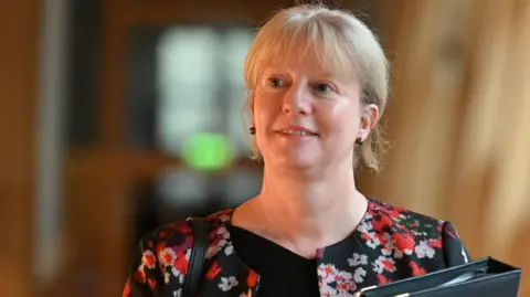 Shona Robison, with blonde hair and wearing a black top and patterned jacket, walks in the Scottish Parliament, carrying a black folder.