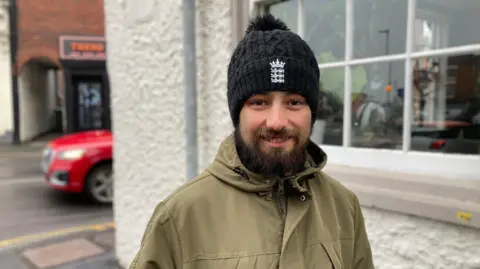 Simon Spark/BBC A young man wearing a black beanie hat with a white logo on the front and a khaki-coloured jacket. He has a dark-coloured beard and moustache and is smiling at the camera 
