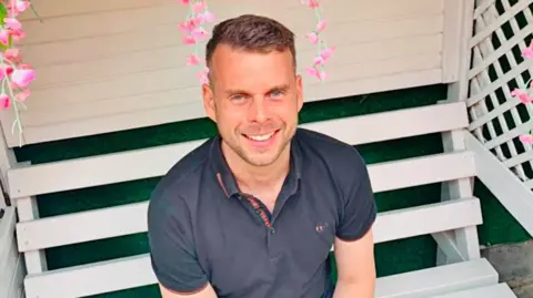 Gavin Campbell is wearing a navy blue polo top while sat on a white bench. He has light stubble and short brown hair.
