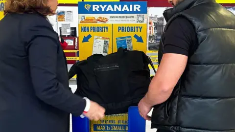 Two unidentified passengers, a man and a woman wearing black, attempt to put a large black bag into a Ryanair bag measurement box. It doesn't look like it fits. The box is in Ryanair's blue and yellow colours, with the words "priority" and "non-priority" with arrows. 