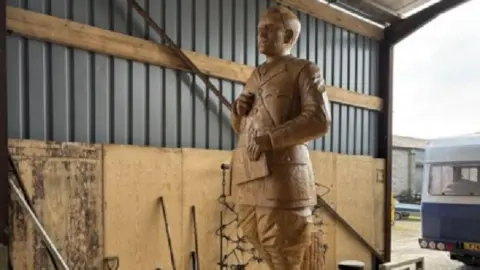 One of the oak statues of JRR Tolkien, in what looks like a barn in the countryside. He is looking serious and is holding a book.