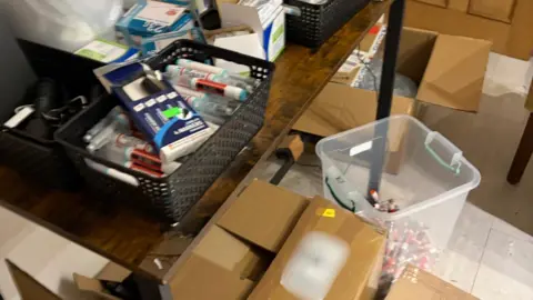 MHRA The image shows a storage room with cardboard boxes, and a clear plastic tub with pre-filled weight-loss pens. On a table there is a black basket with more weight-loss injections