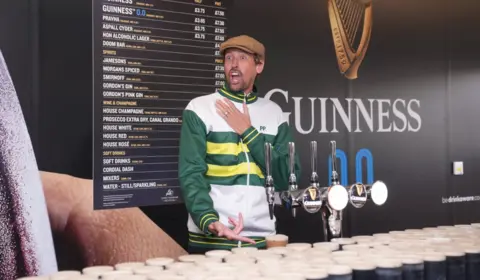 Peter Crouch in a flat tweed cap and wearing a racing jacket with green, yellow and white stripes. He is standing behind a makeshift bar filled with dozens of pints of Guinness crammed onto it and a sign in the background advertising the beer. Crouch is looking fake-shocked with his mouth wide open and pointing to the beer.