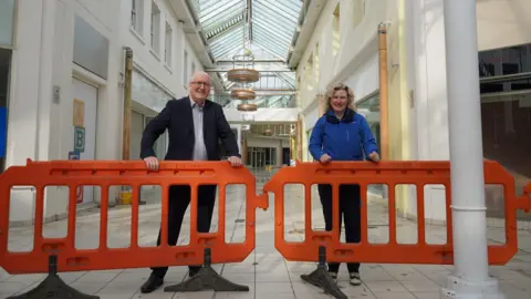 A man and a woman stood infant of a derelict shopping centre infront of construction orange gates