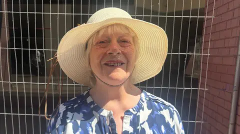 Harvey Bell/BBC A woman in a sun hat and a flowery top stands in front of Heras fencing.
