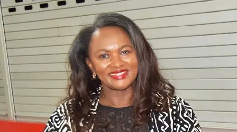 Emertha Uwanyingira is a middle aged Black woman of Rwandan heritage. She has long brown hair and is wearing a black top with a loose black and white print jacket over it. One earring is visible in her right ear. She is wearing red lipstick and smiling at the camera. She is standing in front of a red counter with silver shutters pulled down over it. It looks like a canteen.
