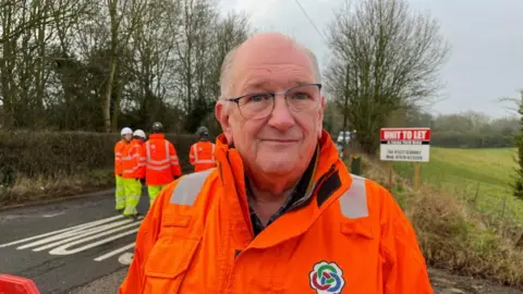 Ollie Conopo/BBC Man wearing glasses and orange hi-viz with road maintenance workers in the background