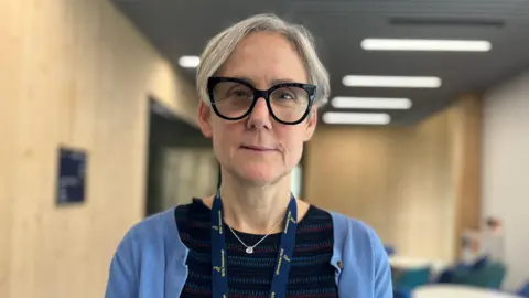 Emma Baugh/BBC Esther Norton looks at the camera. It is a shot of her head and shoulders. She has medium-length grey hair and is wearing glasses with large black frames. She has a light blue cardigan over a stripy, darker-blue top. She is wearing a small silver pendant. The dark blue ribbon of a lanyard, most of which is out of the picture, is around her neck. The background, a lecture room with wood-coloured walls and overhead lights, is out of focus.