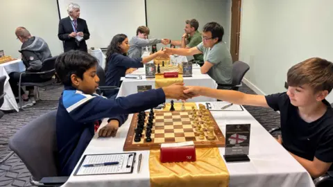 Simon Thake/BBC Chess players face off against each other across a number of square tables in a hotel room. The players shake hands across the table.