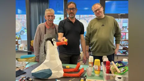 Artworks Art4All CIC Three man stand behind their model seagull which is made of cardboard. It has dark hair and thick eyebrows. It is surrounded by arts and crafts items including paint tubes and pliers.