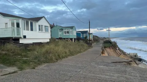 Andrew Turner/BBC Wooden homes set back from concrete roadway which is being undermined by the sea.