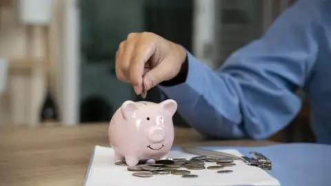 Getty Images A hand putting money into a piggy bank, with lots of coins scattered around it on a table
