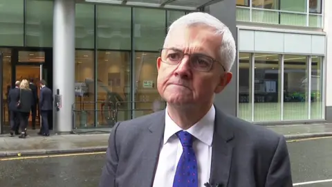 BBC Chris Huhne outside the High Court wearing a suit with a blue tie. He has white hair and glasses