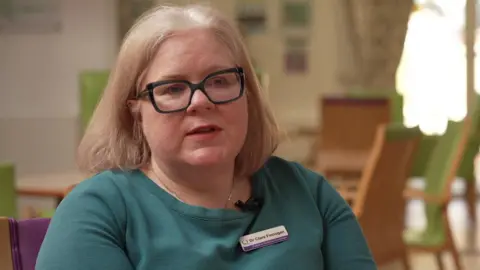 Clare Finnegan has shoulder length light brown hair, is wearing glasses, and a turquoise jumper.