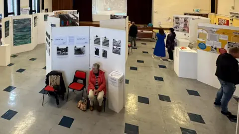 TINA BRENNEN The exhibition seen from above. There are people looking at white boards featuring pictures and information positioned on a black and white tiled floor.