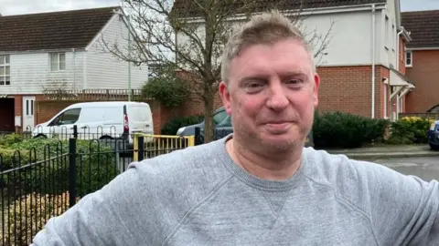 Vikki Irwin/BBC Simon Burton is outside in the street on a housing estate. He is wearing a grey jumper and is smiling at the camera.