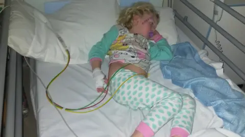 Emma Alderman Noa-Rose sleeping in a hospital bed with various wires coming out of her stomach. She wears peppa pig themed pyjamas and has a dummy in her mouth.