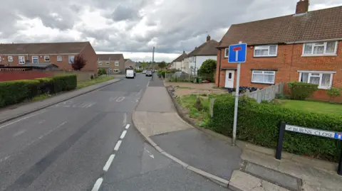 Street view shows red brick houses (left and right) either side of a grey road which has a blue and white T above a street sign for Burns Close