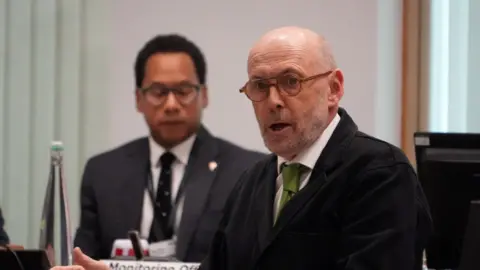 John Fairhall/BBC A man wearing a black jacket over a white shirt and green tie. He also has glasses on and appears to be in the middle of talking. 