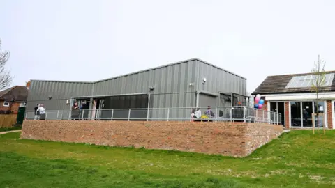 A grey, single storey building on an elevated brick platform. Some baloons can e seen by the side entrance of the property.