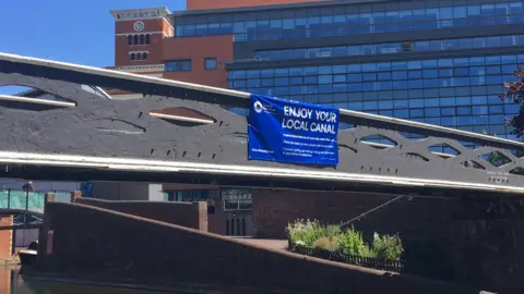 A sign put up above the canal in Birmingham's Brindley Place by the Canal and River Trust encouraging people to 'enjoy your canal' but give others space