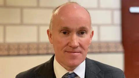 Laura Coffey/BBC MAn with very short grey hair wearing a grey jacket and lanyard