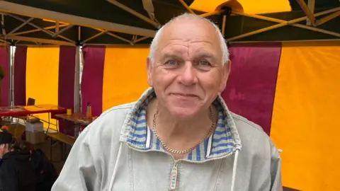 A man stood smiling at the camera wearing a beige zipped jumper, backed by purple and yellow marquees.