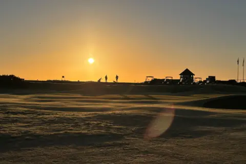 Peter Mulvey Golfers silhouetted on the course against an orange sunrise glow.