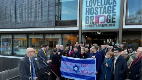 Tom Grundy/BBC People together holding a flag with 'Bring them home now' written on it.