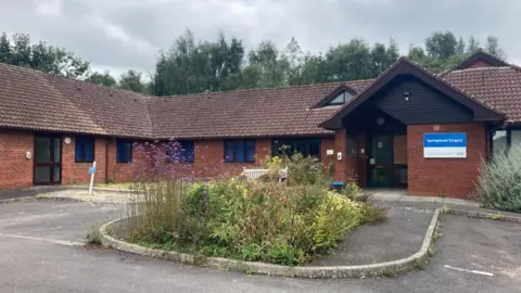 LDRS An image of the currently disused Springmead Surgery which closed two years ago but will take in patients from April 14th. A red brick single storey building with an overgrown front garden and car park out the front.