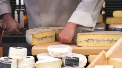 A picture of a cheese counter. A big wheel of hard aged cheese is being cut by a cheese-monger in white. 