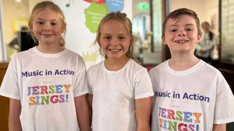 Hayley, Flo and Oak are standing next to each other. They are all smiling and wearing white T-shirts, two of which have Jersey Sings! on them.