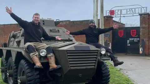 Nick Taylor A Saracen armoured personnel carrier parked in front of Fort Paull. The six-wheeled vehicle, which is a brown/green colour, is parked next to a green corrugated metal building. Nick Taylor and Nick Moore are both sat on the vehicle holding their arms out.