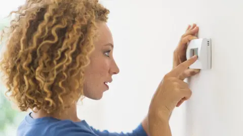 Getty Images A woman operates a thermostat on the interior wall of a house