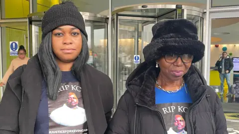 PA Media Tellecia Strachen and Wendy Clarke, wearing black coats, black hats and T-shirts with RIP KC and a picture of Mr Clarke printed on. They are standing outside the building in which the hearing was held and both look unhappy but resigned.