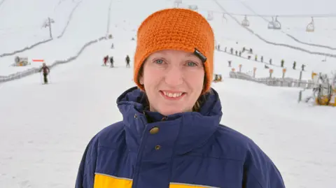Annette Cockburn, wearing an orange knitted hat and a blue winter jacket, stands on a snowy ski slope, with chairlifts, skiers, and ski infrastructure visible in the background.