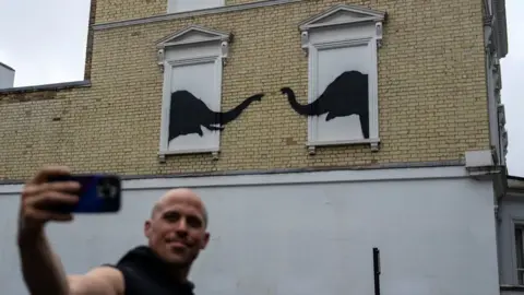 Getty Images A man takes a selfie in front of a Banksy artwork in London. The artwork shows two elephants painted over windows, their trunks reaching towards each other