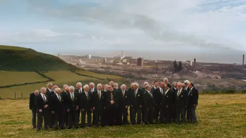 Roo Lewis Men in club blazers and ties stood on a hill with Port Talbot steelworks in the backdrop